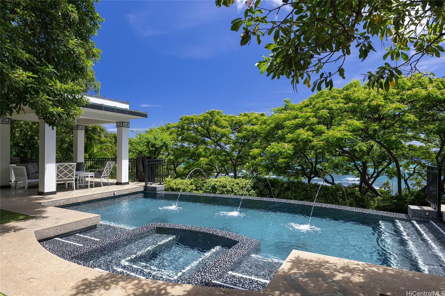 This image showcases a luxurious pool and spa area with a stunning ocean view. The pool features a dark-tiled spa section with steps, and fountains add a touch of elegance. A covered patio with seating is visible on the left, and lush greenery frames the scene, creating a serene and inviting atmosphere.