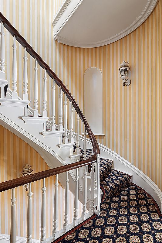 This image showcases a beautifully designed staircase with a curved railing and white balusters. The walls are adorned with striped wallpaper, and a patterned carpet covers the stairs, adding a touch of elegance. The overall impression is one of classic sophistication and attention to detail.