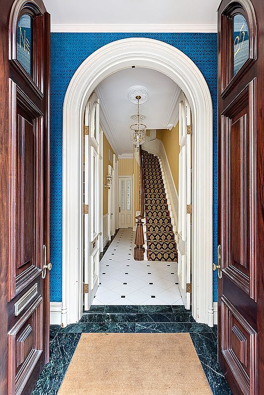 The image showcases an elegant entryway and hallway, framed by open wooden doors. The hallway features a striking blue patterned wallpaper, a white arched doorway, and a staircase with a patterned runner. The flooring transitions from a dark green marble at the entrance to white tiles with black accents further into the hallway, creating a sophisticated and inviting atmosphere.