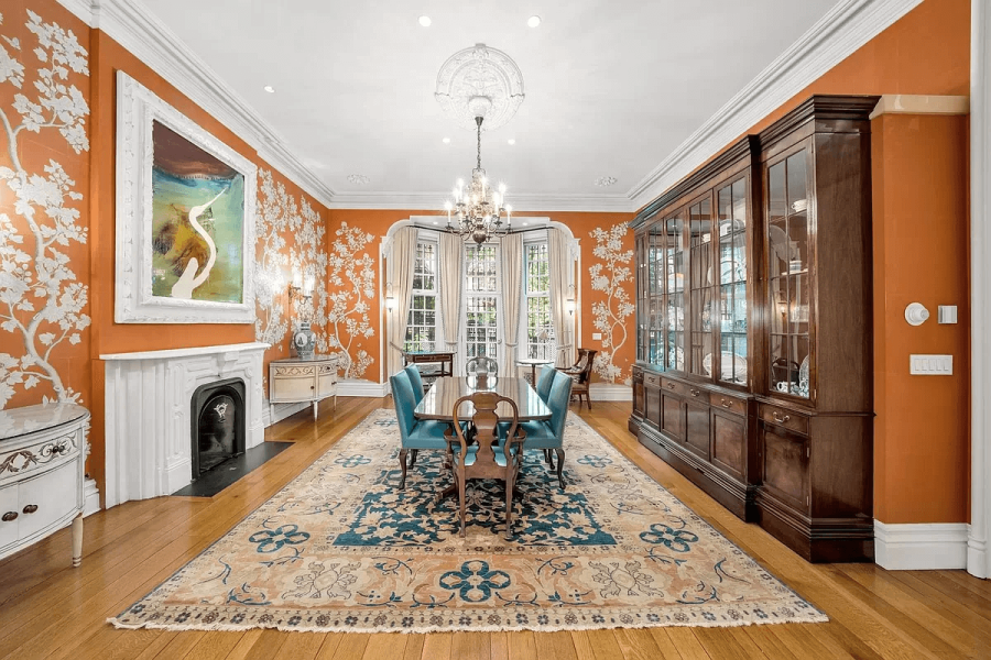 This interior shot showcases a formal dining room with hardwood flooring and a large oriental rug. The walls are painted in a striking orange hue with a floral pattern, complemented by white trim and a built-in wooden china cabinet. A chandelier hangs above a dining table surrounded by blue upholstered chairs, creating an elegant and inviting atmosphere.