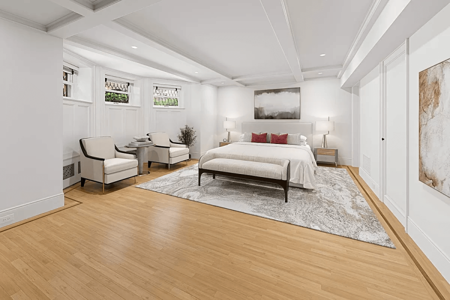 This is a bright and spacious primary bedroom featuring a light color palette. The room includes a king-size bed with a bench at the foot, two armchairs near the windows, and a large area rug that adds texture and visual interest. The hardwood floors and coffered ceiling contribute to a sophisticated and comfortable living space.