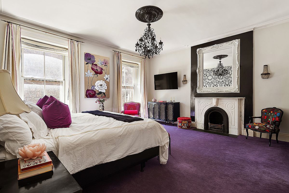 This is a luxurious primary bedroom featuring a large bed with plush pillows and a white duvet. The room is decorated with a black chandelier, a decorative fireplace with a large mirror above it, and a flat-screen TV. The purple carpet adds a touch of elegance and warmth to the space.