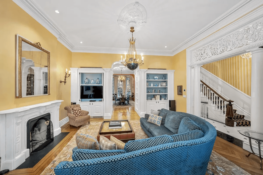 This is a luxurious living room featuring yellow walls, white trim, and hardwood floors. The room has built-in bookshelves, a fireplace, and a large, curved, blue sofa. The overall aesthetic is elegant and sophisticated, suitable for a high-end real estate listing.