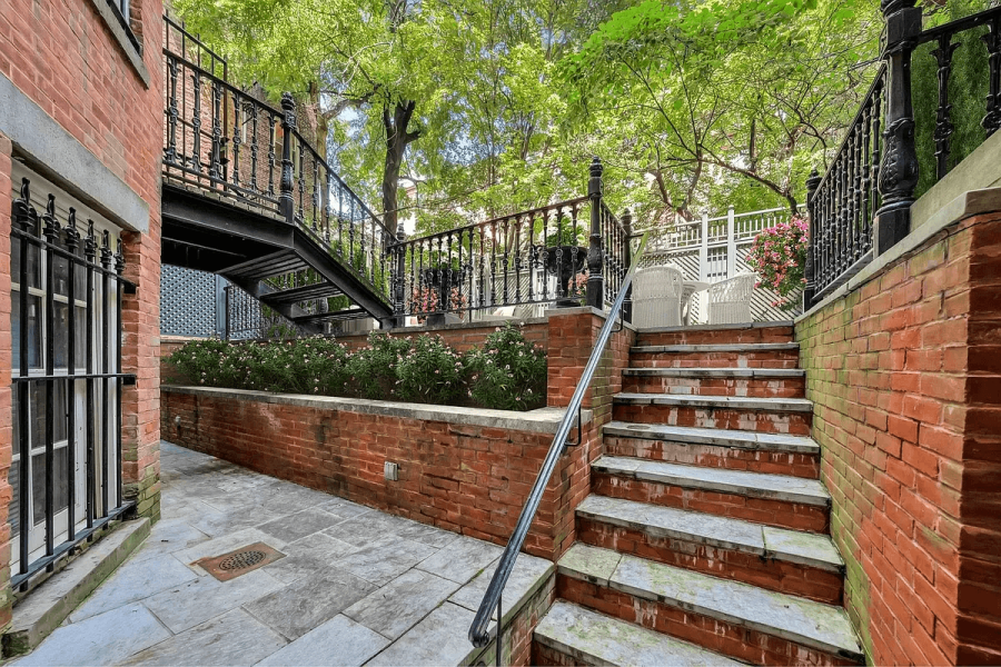 This exterior shot showcases a charming yard or garden area. Brick walls and stone steps create a tiered effect, leading to an upper level. A black iron staircase adds architectural interest, while lush greenery and flowering plants enhance the overall appeal of the space.