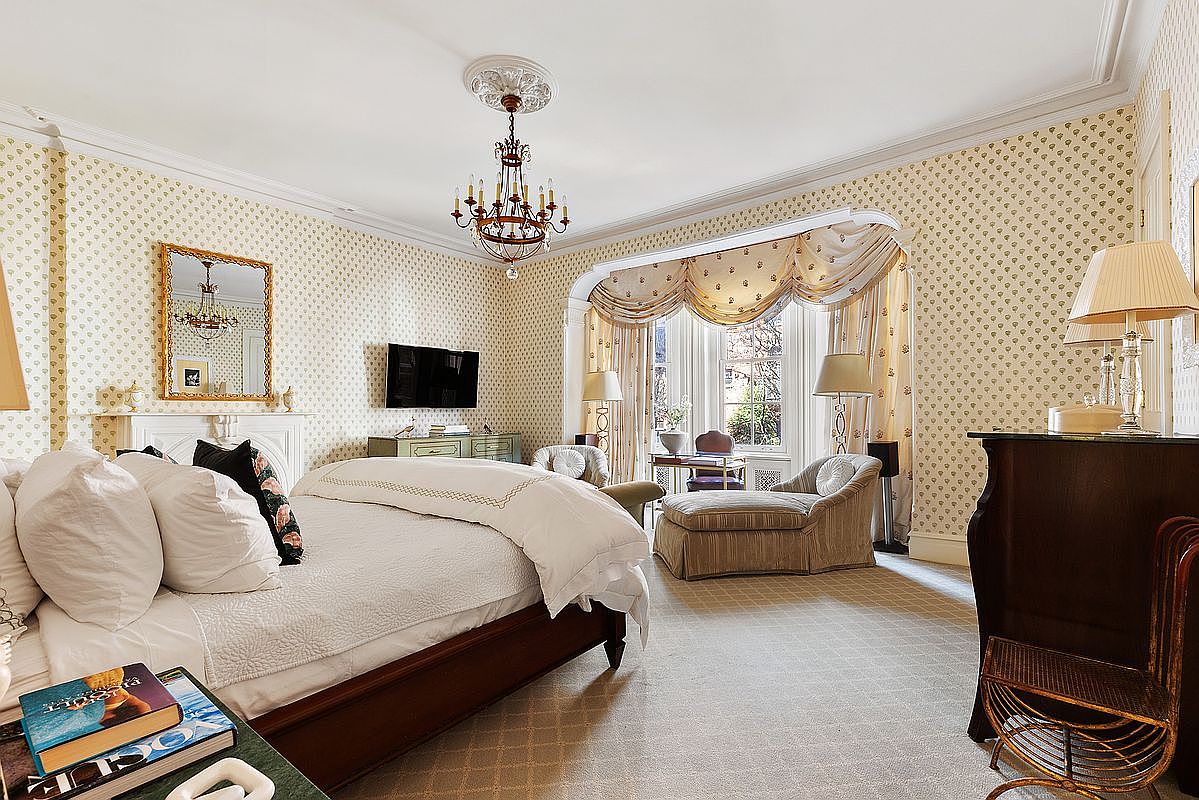 This is a luxurious primary bedroom featuring patterned wallpaper, a chandelier, and a large window with elegant drapery. The room includes a bed with white linens, a fireplace with a mirror above, and a chaise lounge near the window, creating a comfortable and sophisticated atmosphere. The perspective is from the foot of the bed, showcasing the room's depth and features.
