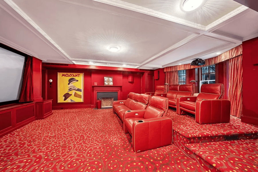 This is a home theater room with red walls and a red patterned carpet. The room features tiered seating with red leather theater chairs and a screen on the left wall. A yellow and black poster hangs over a fireplace.