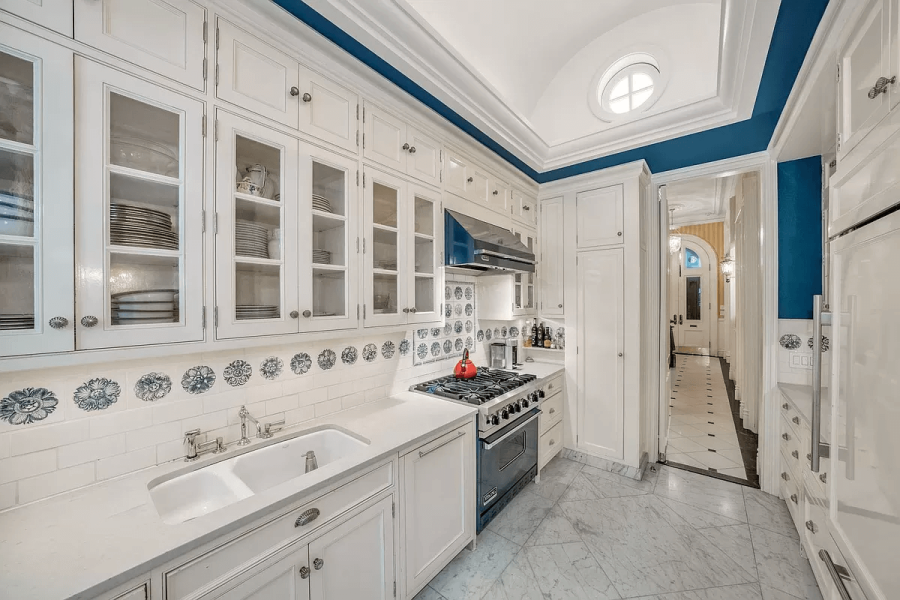 This is a bright and elegant kitchen featuring white cabinets, glass-front displays, and a striking blue accent trim. The kitchen is equipped with modern appliances and offers ample counter space. The marble flooring adds a touch of luxury and sophistication to this space.