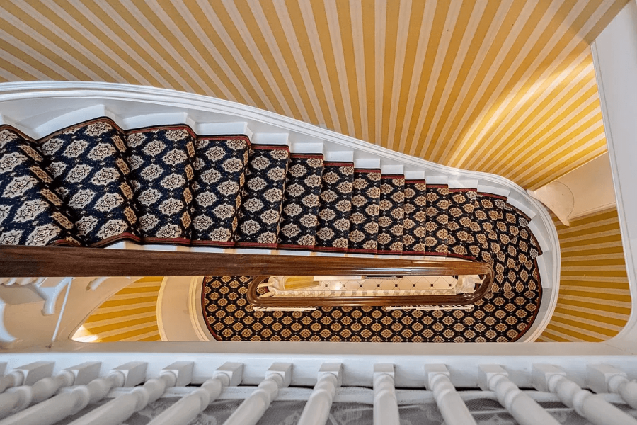 This is an overhead view of a winding staircase. The stairs are carpeted with a patterned rug, and the walls are decorated with yellow and white stripes. The banister is white with a wooden handrail, creating a classic and elegant impression conducive to high-end property appeal.