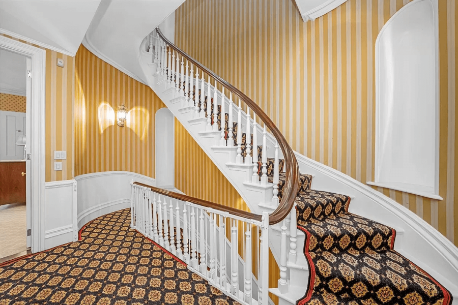 This image depicts an elegant interior hallway featuring a curved staircase. The walls are adorned with yellow and white striped wallpaper, complementing the white-painted staircase with a wooden handrail. A patterned runner covers the stairs and hallway, adding a touch of classic style to the space.
