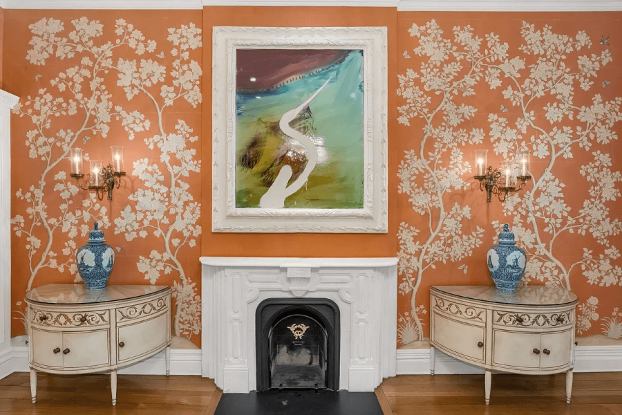 This interior shot showcases a meticulously decorated living room, instantly conveying elegance and refined taste. The room features a fireplace with a white mantle, flanked by matching side tables and accent lamps. The orange wallpaper with painted trees provides a unique and sophisticated backdrop, complementing the artwork above the fireplace.