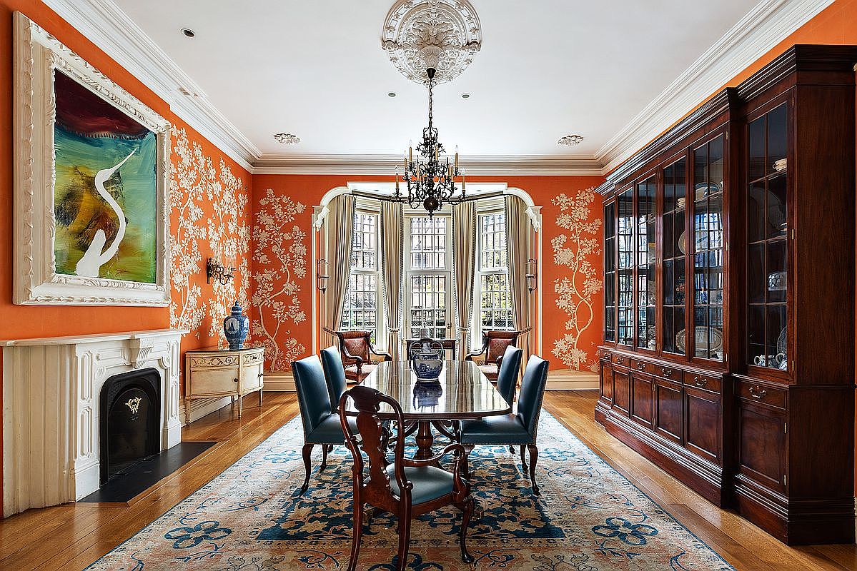 This is an elegant dining room featuring orange walls with white floral patterns, a large wooden dining table with blue upholstered chairs, and a striking chandelier. A built-in wooden cabinet stands against one wall, while a fireplace with a white mantel and a framed painting adorn another. The room is illuminated by natural light streaming through a bay window, creating a sophisticated and inviting atmosphere.