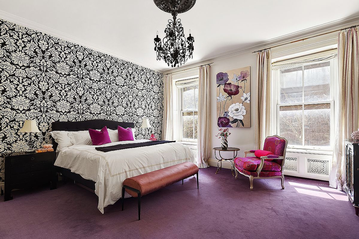 This is a luxurious primary bedroom featuring a black and white damask wallpaper accent wall, a black upholstered bed with white linens and purple pillows, and a striking black chandelier. The room is carpeted in purple and includes a decorative chair, a floral painting, and large windows with elegant curtains, creating a sophisticated and opulent atmosphere. The perspective is from the foot of the bed, capturing the entire room.