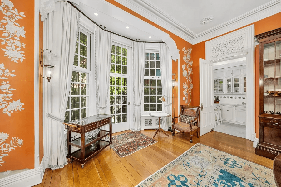 The image showcases a beautifully decorated living room with hardwood floors and orange walls adorned with white floral accents. A bay window provides ample natural light, complemented by elegant curtains and sophisticated furnishings. The room exudes a classic and refined ambiance, perfect for entertaining or relaxing.