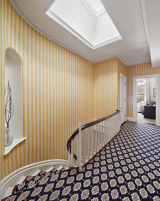 This interior shot showcases a hallway and staircase featuring striped wallpaper in yellow and white, a patterned blue and beige carpet, and a white banister with a wooden handrail. A skylight provides natural light, and an arched niche displays a decorative vase. The overall impression is classic and elegant.