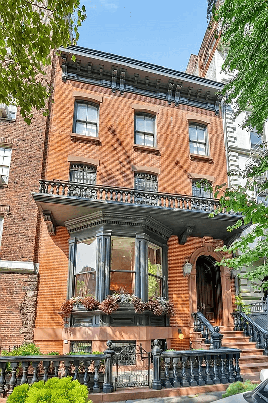 This is a full frontal view of a classically designed brick townhouse. The building features a prominent bay window on the second level, iron railings on the balcony, and an elegant arched entryway. The brickwork, paired with the dark trim, gives the property a distinguished and stately appearance.