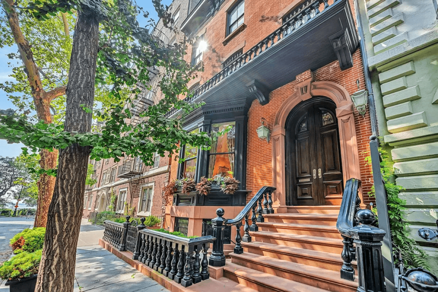 The image showcases the entryway of a beautiful brick townhouse. A grand, arched doorway is accessed by a set of stairs with ornate black railings. The building features a bay window with flower boxes and a decorative balcony, enhancing the overall curb appeal and creating a welcoming first impression.