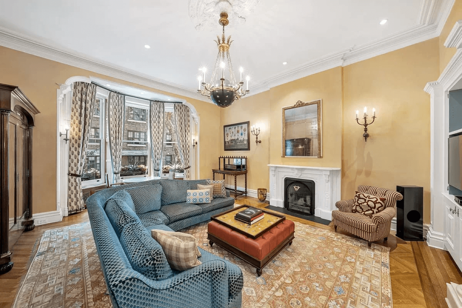 This is a traditionally styled living room featuring a blue patterned sofa and armchair, with a red ottoman serving as a coffee table. The room boasts a decorative fireplace, ornate light fixtures, and a bay window with patterned curtains. A large area rug grounds the space, creating a warm and inviting atmosphere.