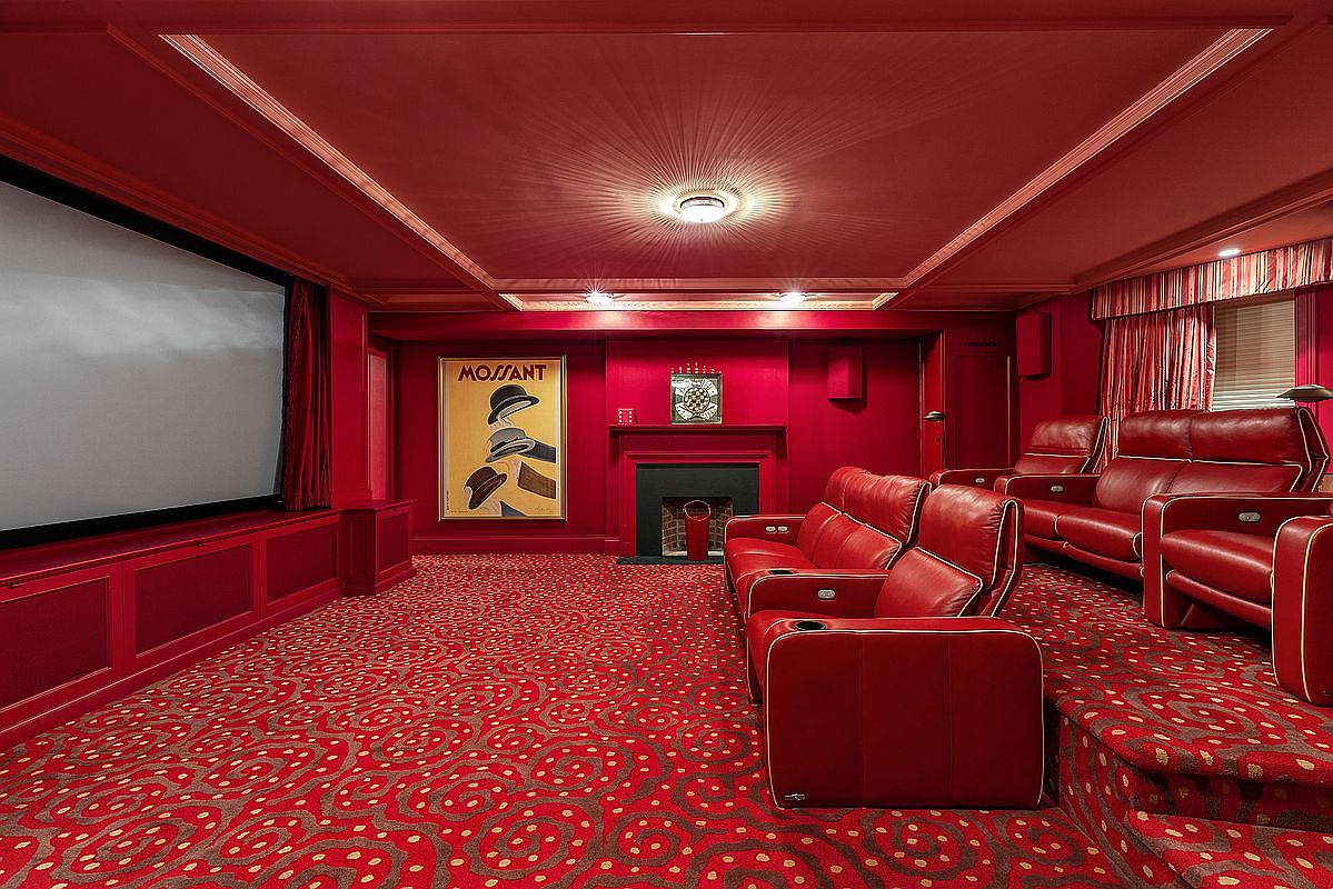 This is an interior shot of a home theater room, featuring red walls, a patterned red carpet, and red leather reclining chairs with white trim. A large screen is visible on the left, and a fireplace with a decorative clock above it is centered on the back wall, with a vintage poster to the left of the fireplace. The room is designed for a luxurious viewing experience.