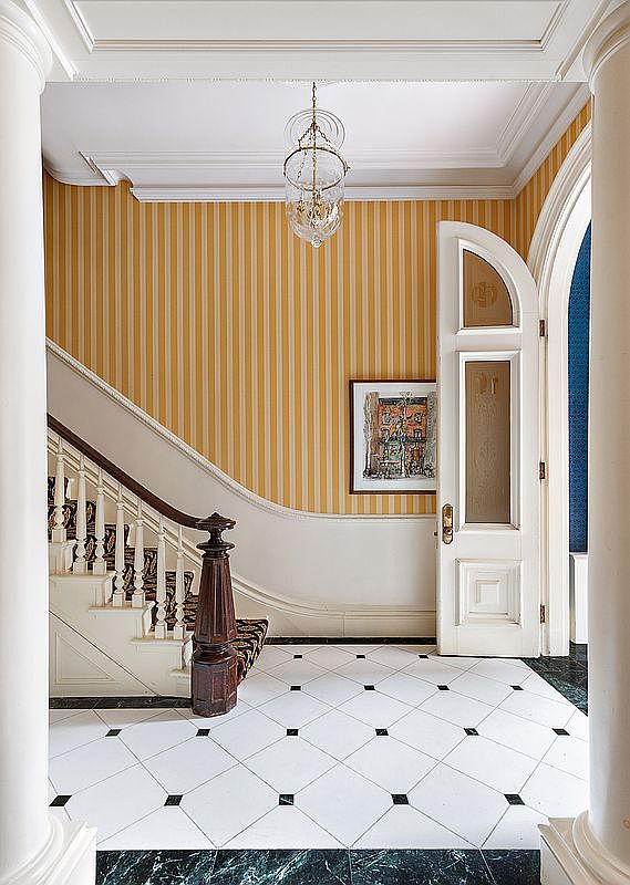 This interior shot showcases a grand entryway with a staircase. The space features yellow striped wallpaper, a white staircase with dark wood accents, and a white tile floor with black accents. A chandelier hangs from the ceiling, and a partially open door reveals a glimpse of another room, creating an inviting and elegant atmosphere.