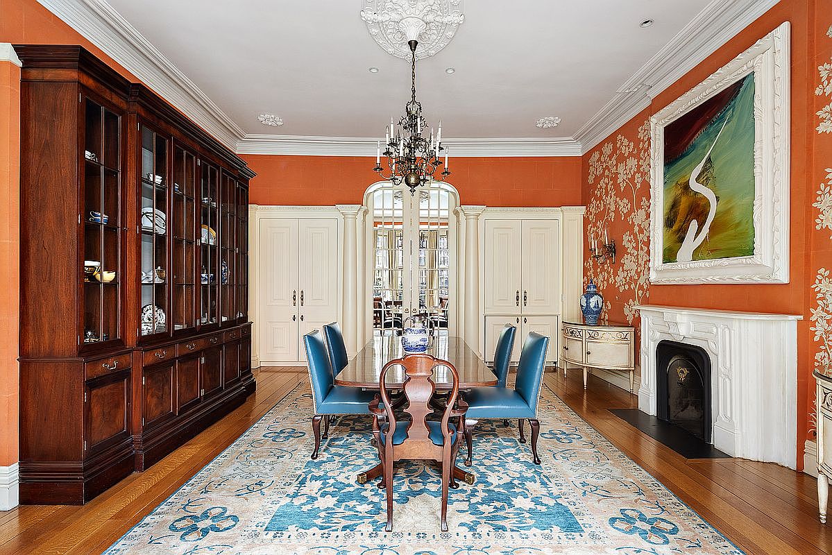 This is a formal dining room featuring a large wooden table surrounded by blue upholstered chairs. A grand wooden cabinet stands against one wall, while a fireplace and artwork adorn another. The room is decorated in a traditional style with orange walls, a patterned rug, and an ornate chandelier, creating an elegant and inviting atmosphere.