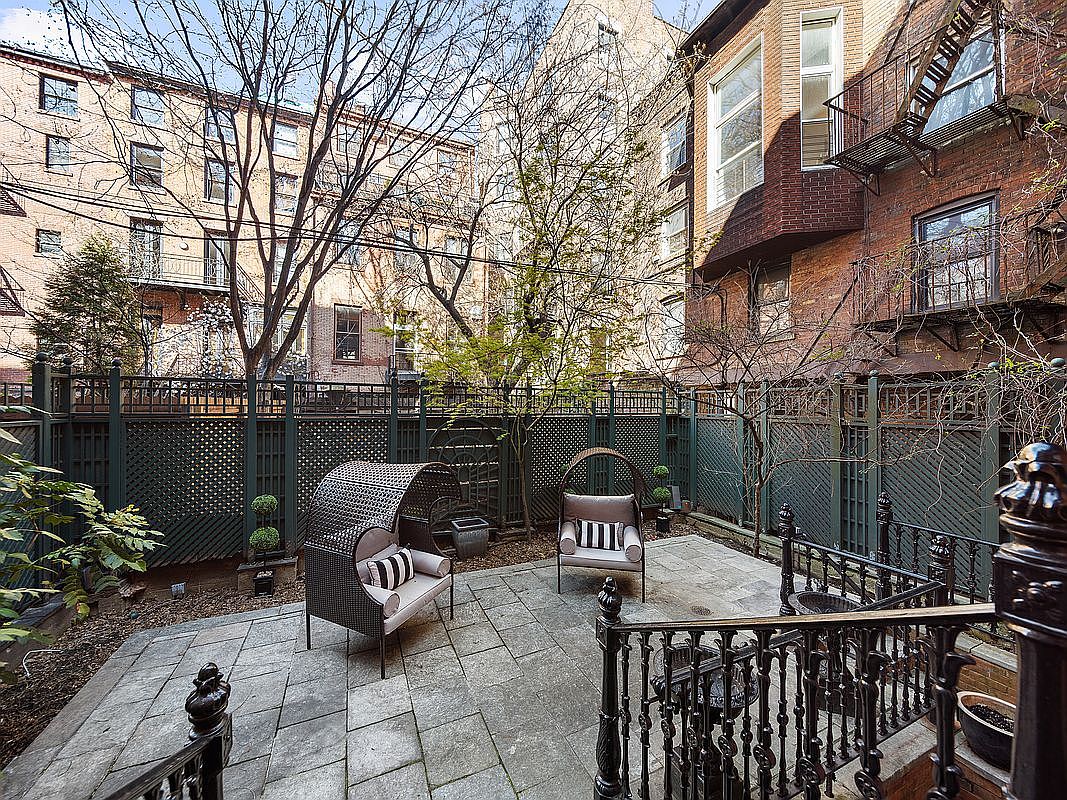 This image showcases a charming backyard garden area, featuring a stone patio with stylish outdoor seating. A dark green lattice fence provides privacy, while mature trees and greenery add to the serene atmosphere. The space is well-maintained and offers a relaxing outdoor retreat.