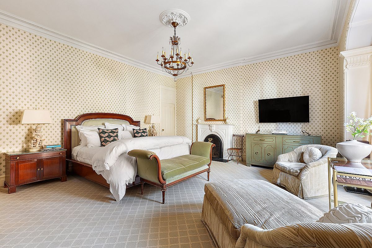 This is a primary bedroom featuring a large bed with a wooden headboard, a chandelier, and a fireplace. The walls are covered in patterned wallpaper, and there is a TV mounted on the wall. The room is furnished with a variety of seating options, including a chaise lounge and an armchair, creating a luxurious and comfortable atmosphere.