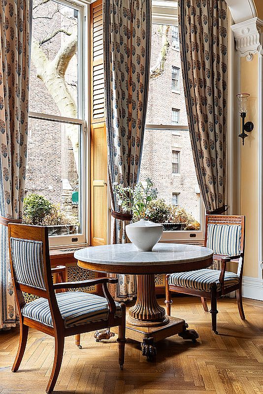 This is an interior shot of a dining room featuring a round marble-top table set between two striped upholstered chairs. The room is bathed in natural light from large windows adorned with patterned curtains, offering a view of the exterior brick buildings and greenery. The herringbone wood flooring adds a touch of classic elegance to the space.