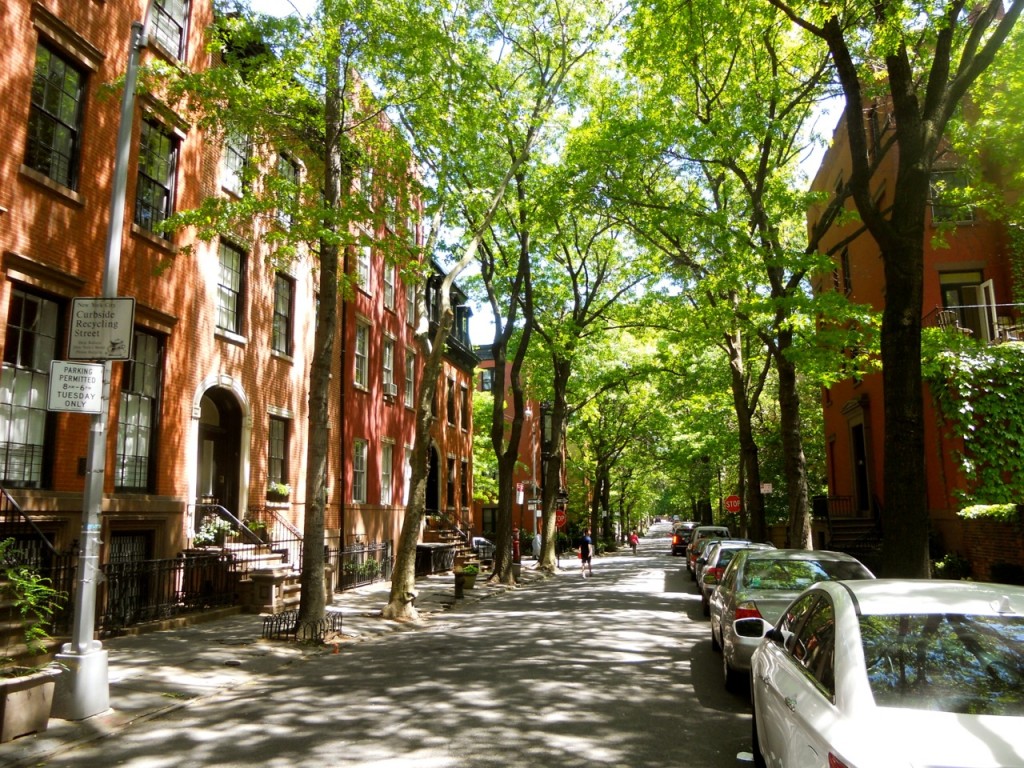 The image showcases a charming residential street lined with brick townhouses. Mature trees create a shaded canopy over the road, enhancing the neighborhood's appeal. Parked cars suggest convenient street parking, and the well-maintained facades contribute to the overall inviting atmosphere.