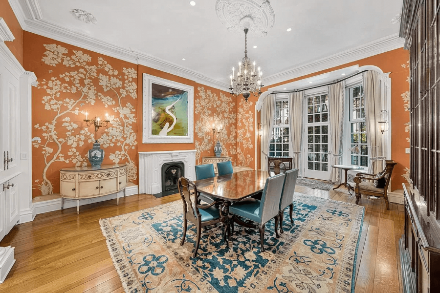 The image showcases an elegant dining room with orange floral wallpaper, a wooden dining table with blue upholstered chairs, and a patterned area rug. Natural light streams in through a bay window, complemented by a chandelier overhead. A fireplace with a decorative mantel and a sideboard enhance the room's classic charm and sophistication.