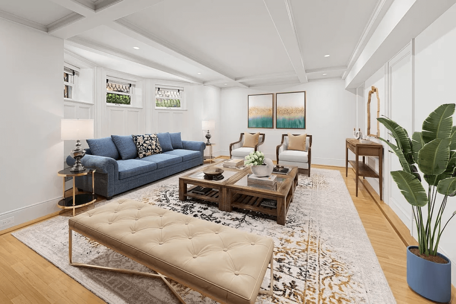 The living room features a blue sofa adorned with decorative pillows, centered around a wooden coffee table atop a patterned area rug. Two armchairs face the couch, flanking two pieces of art. Natural light filters in through the window and the room is decorated with a number of interior design pieces.