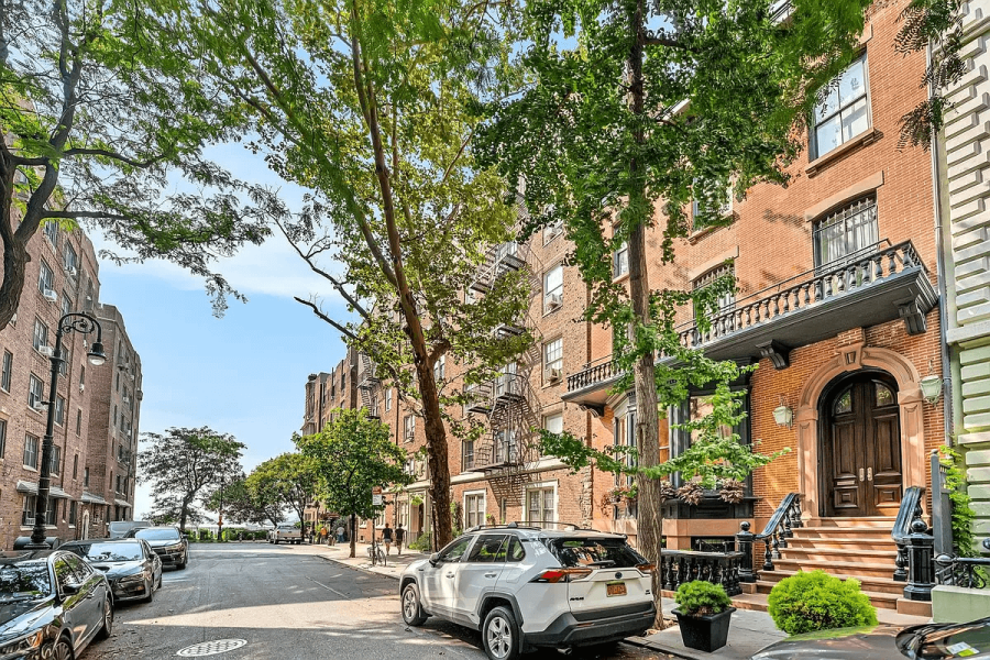The image shows the front of a classic apartment building with brick facade and stone detailing. Lush green trees line the street, adding to the curb appeal. Several cars are parked on the street, suggesting a vibrant urban environment.