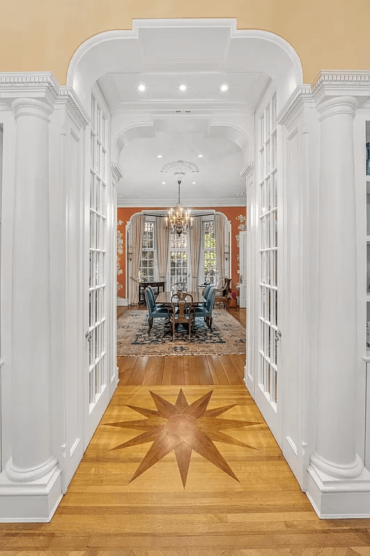 This interior shot highlights a formal dining room as seen through an arched entryway. The blonde wood floor features a striking starburst inlay. The dining room features a neutral rug, burnt orange walls, bay window, and crystal chandelier.