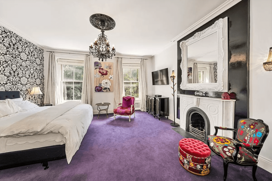 This primary bedroom features a unique design with a decorative black and white wallpaper on one wall and a bold purple carpet. A fireplace with a white mantel and a large decorative mirror serves as a focal point on the opposite wall. The room is furnished with a bed, accent chairs, a television, and a statement chandelier, creating a luxurious and visually striking atmosphere.