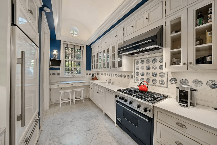 This is a well-lit kitchen featuring elegant white cabinetry with glass-front upper cabinets complemented by a black range hood. The blue accent wall adds a touch of sophistication, while the tile backsplash and marble flooring enhance the upscale aesthetic. The kitchen appears clean and organized, ready for culinary endeavors.