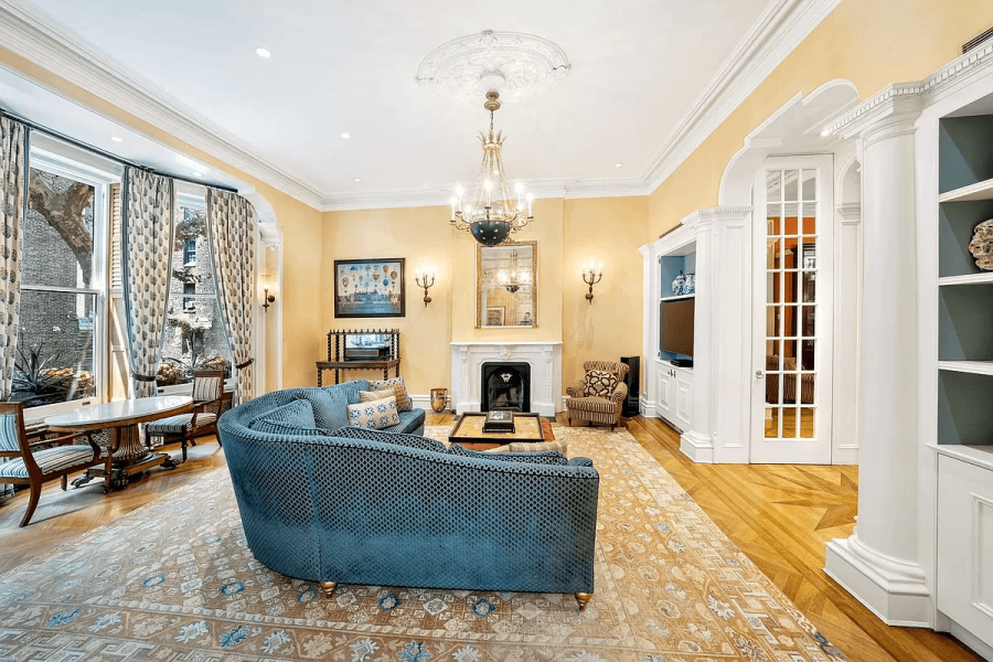This elegant living room features high ceilings, intricate crown molding, and a stunning chandelier. A curved blue sofa anchors the space, complemented by a patterned area rug and hardwood flooring. The room also includes a fireplace, built-in shelving, and a window with draperies, creating a sophisticated and inviting atmosphere.