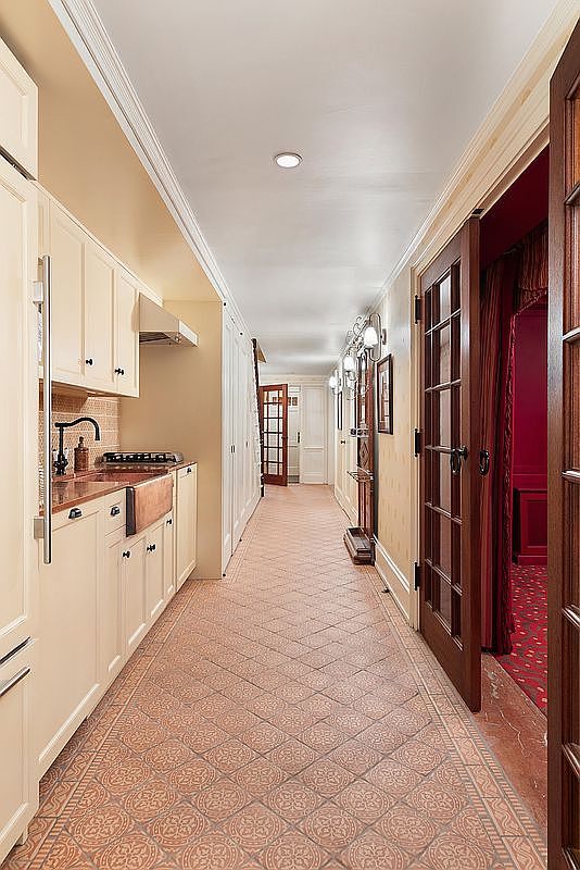 This interior shot showcases a long hallway featuring a unique patterned tile floor and a combination of kitchen elements on the left and paneled doors on the right. The hallway is well-lit with overhead lighting and wall sconces, leading to other rooms in the house. The perspective gives a sense of depth and the layout suggests a functional and stylish home.