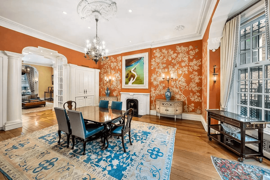 This elegant dining room showcases a blend of classic and ornate design elements, featuring rich orange walls adorned with a tree-branch pattern and a striking blue-and-cream area rug that defines the space. The room is furnished with a dark wood dining table and upholstered chairs, complemented by antique-style sconces and a chandelier adding a touch of vintage charm to the luxurious ambiance.