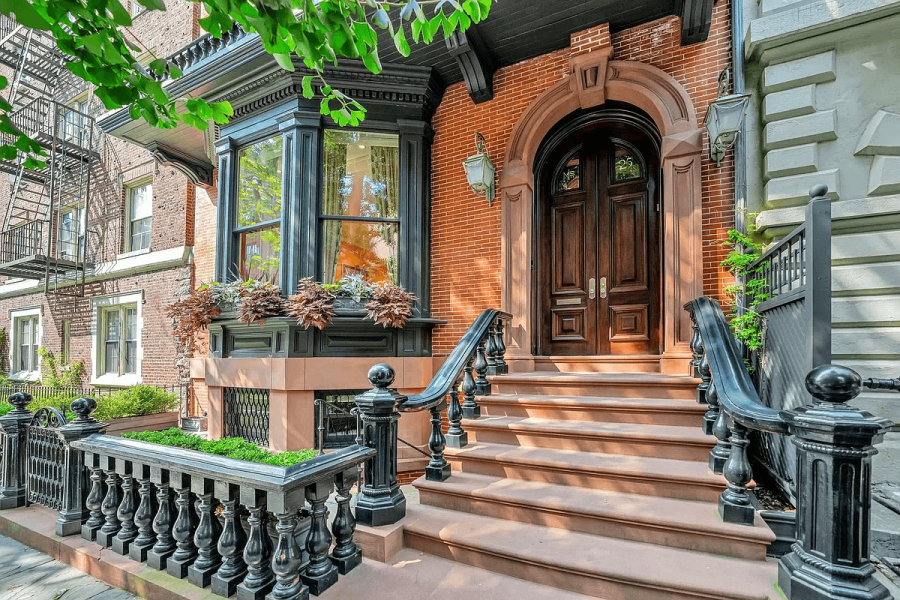 This image showcases the entryway of a charming brick townhouse. A grand wooden door with an arched frame is accessed via tiered steps with elegant black railings. A bay window to the left adds architectural interest, and lush planters provide a touch of greenery, contributing to a welcoming facade.