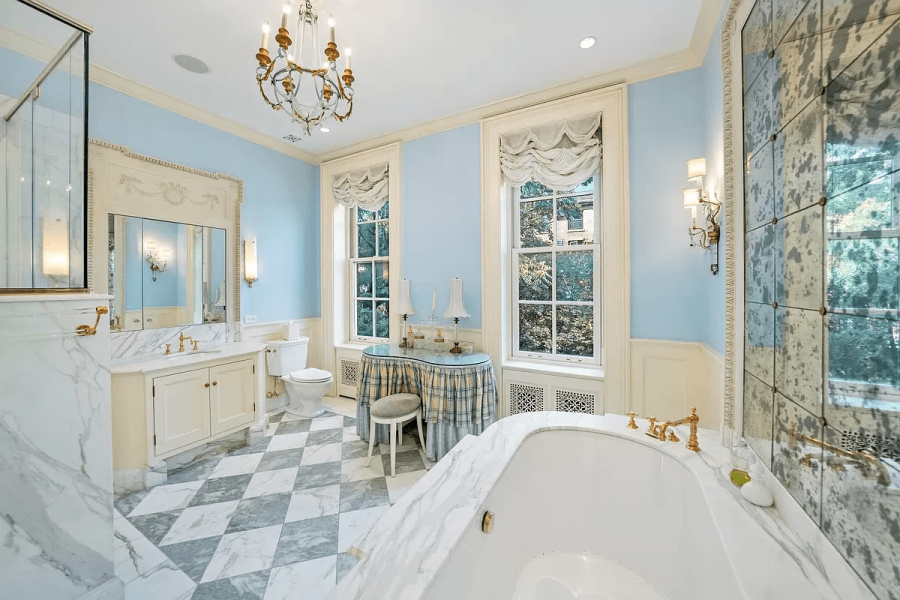 This is a luxurious primary bathroom featuring a marble-topped vanity, a freestanding soaking tub, and elegant light fixtures. The walls are painted a soft blue, and the floor has a checkerboard pattern. The room has a classic and sophisticated style, well-suited for a high-end real estate listing.
