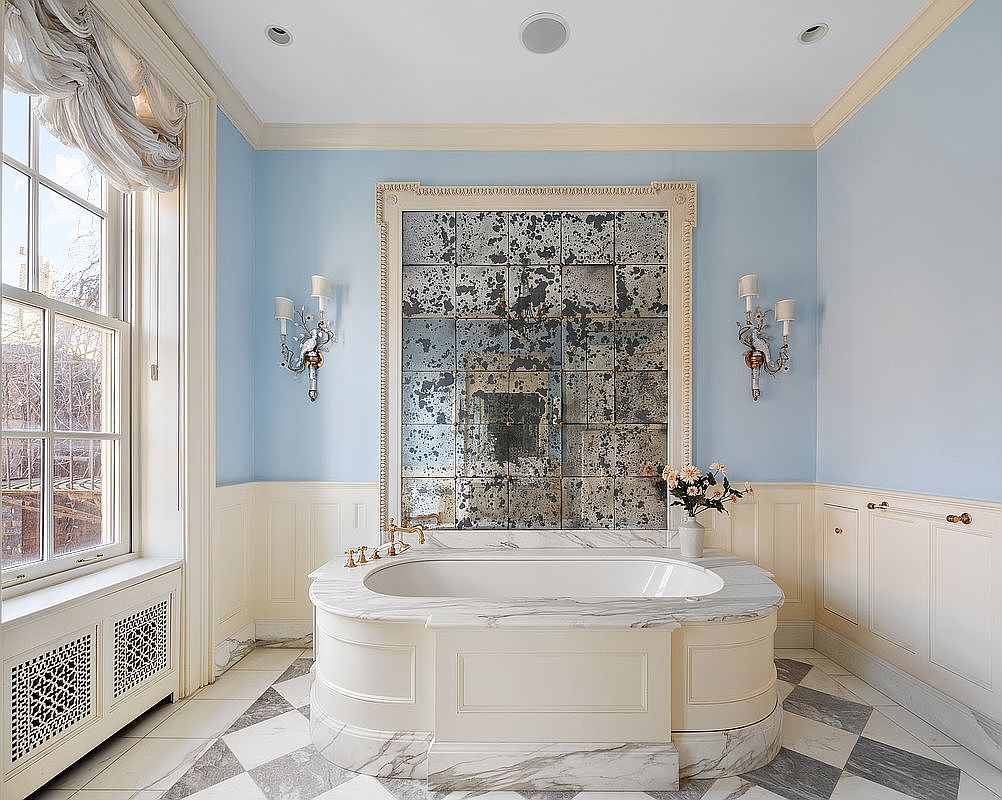 This is a luxurious primary bathroom featuring a freestanding oval bathtub with marble surround, set against a backdrop of a large, antique-style mirrored wall. The room is painted in a soft blue, complemented by white wainscoting and cabinetry, creating a serene and elegant atmosphere. The checkered marble floor adds a touch of classic sophistication.