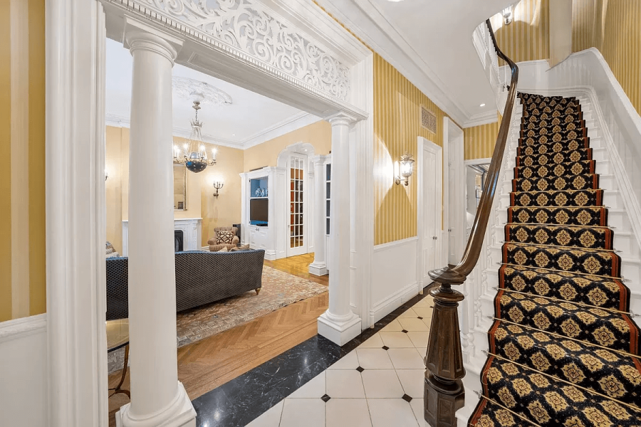 This interior shot captures an elegant hallway featuring a staircase with a patterned runner. The hallway showcases classic architectural details like white columns and decorative molding. Yellow striped wallpaper adds visual interest to the upper walls while the flooring is a mix of marble and wood.