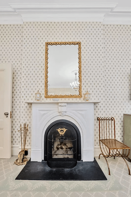 This image showcases a living room featuring a classic fireplace adorned with a decorative mirror and antique accents. The room is wallpapered with a subtle pattern, complementing the overall vintage aesthetic. A unique iron chair adds character, contributing to a cozy and elegant ambiance.