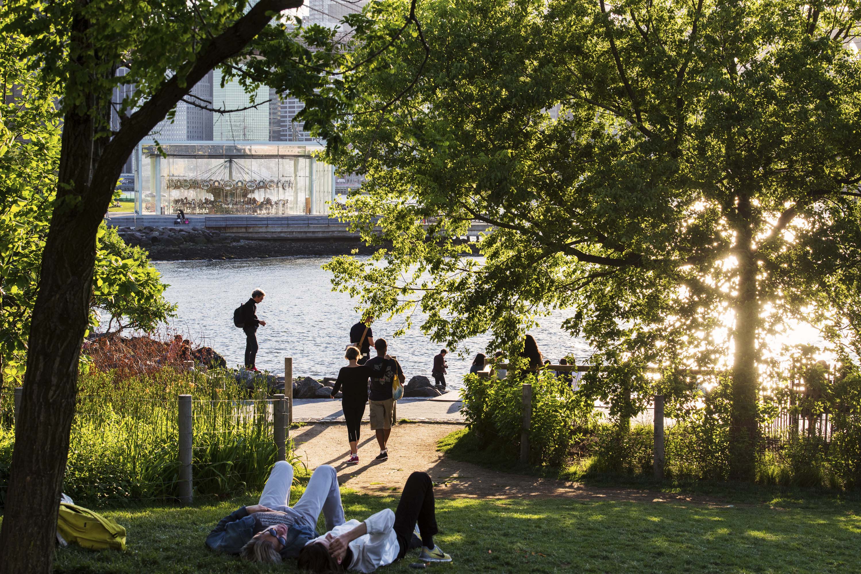 This image showcases a vibrant waterfront park setting. People are enjoying the green space, with some relaxing on the grass and others strolling along the paved pathway towards the water. In the backdrop, a carousel is visible, adding to the park's appeal and making it a desirable recreational area.