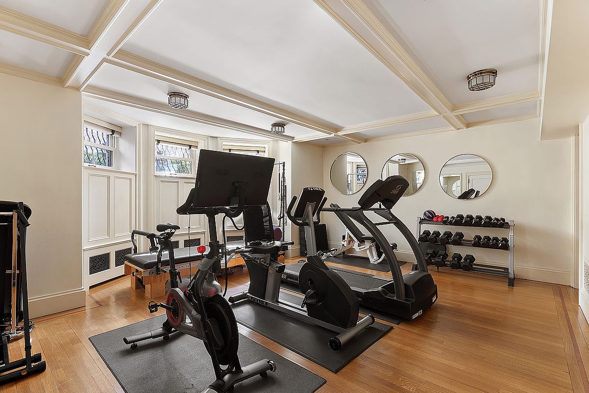This is an interior shot of a well-equipped home gym. The room features hardwood floors, recessed lighting, and three round mirrors on the wall. Exercise equipment includes a stationary bike, elliptical machine, treadmill, and a rack of dumbbells, creating a dedicated space for fitness within the home.
