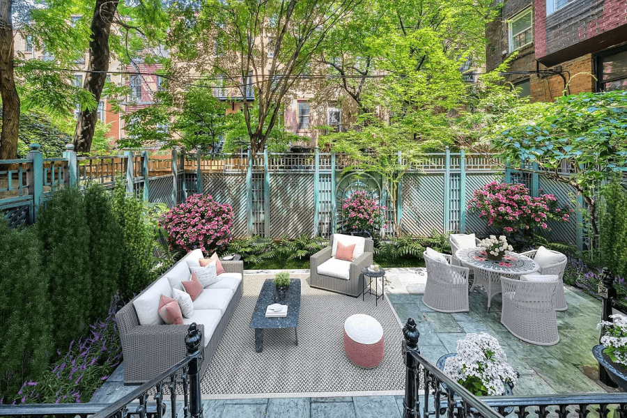 This image showcases a well-maintained backyard garden, designed for relaxation and outdoor living. The space features a patio area with comfortable seating, including a sofa, chairs, and a dining table set, all surrounded by lush greenery and blooming flowers. A decorative fence adds privacy and architectural interest, creating an inviting and serene outdoor environment.