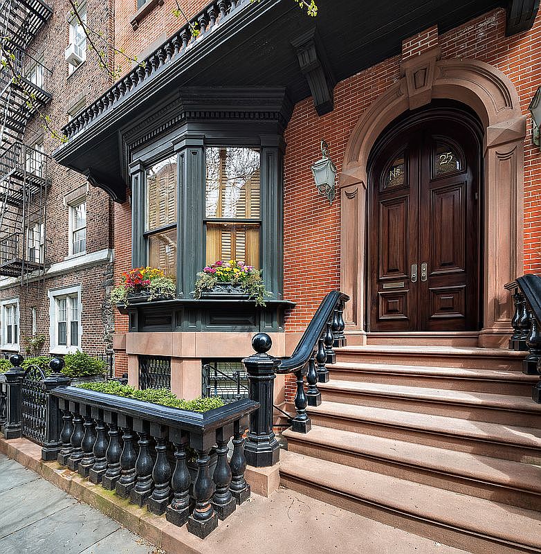 This is a front view of a classic brownstone entryway. The facade features a brick exterior, a dark wood double door with an arched frame, and a bay window with flower boxes. A set of brownstone steps leads up to the entrance, flanked by a black wrought iron railing, creating a welcoming and elegant approach.