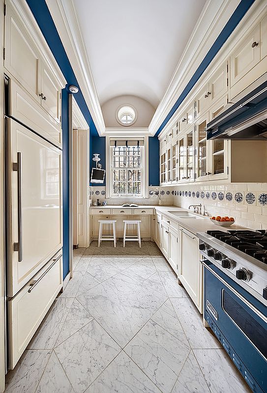 This is a galley-style kitchen with white cabinetry, blue accents, and marble tile flooring. The kitchen features a blue Viking range, a built-in refrigerator, and a small desk area with stools at the end of the room. The overall impression is elegant and well-maintained.