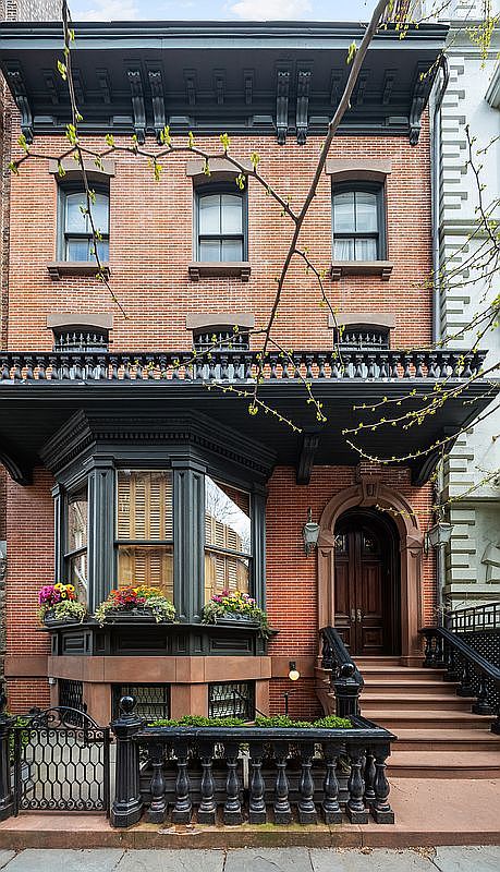 This is a front view of a classic brownstone building. The facade features red brick, dark trim, and a bay window with flower boxes. A set of stairs leads to the front door, and a wrought-iron fence encloses the front yard, creating a welcoming and elegant entrance.