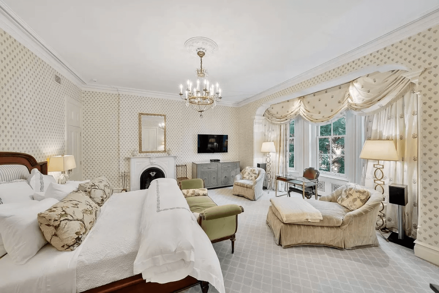 This is a primary bedroom interior, showcasing a luxurious and spacious design. It features a large bed, a seating area with an armchair and a chaise lounge, a fireplace with a decorative mirror above, and a flat-screen TV. The room is decorated with patterned wallpaper, elegant curtains, and a chandelier, creating a sophisticated and comfortable ambiance.