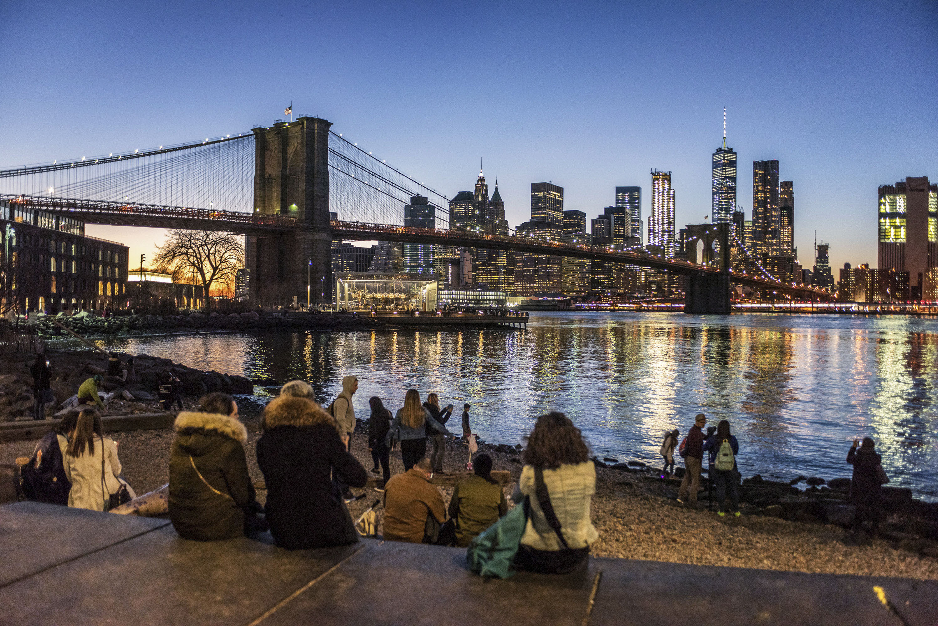 This photograph showcases a vibrant waterfront scene at dusk, featuring the iconic Brooklyn Bridge and illuminated Manhattan skyline. The waterfront area appears to be a popular location, with several people enjoying the views, contributing to the sense of community and offering a lively outdoor space. The combination of the bridge, skyline and waterfront amenities suggest a desirable location for residents.
